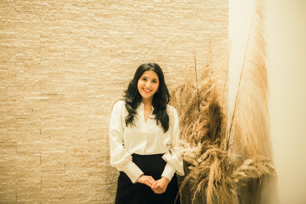 Dr. Zainab Aziz, board-eligible endodontist, smiling in a professional setting with a textured wall and decorative plants, representing advanced endodontic care and patient-centered philosophy.